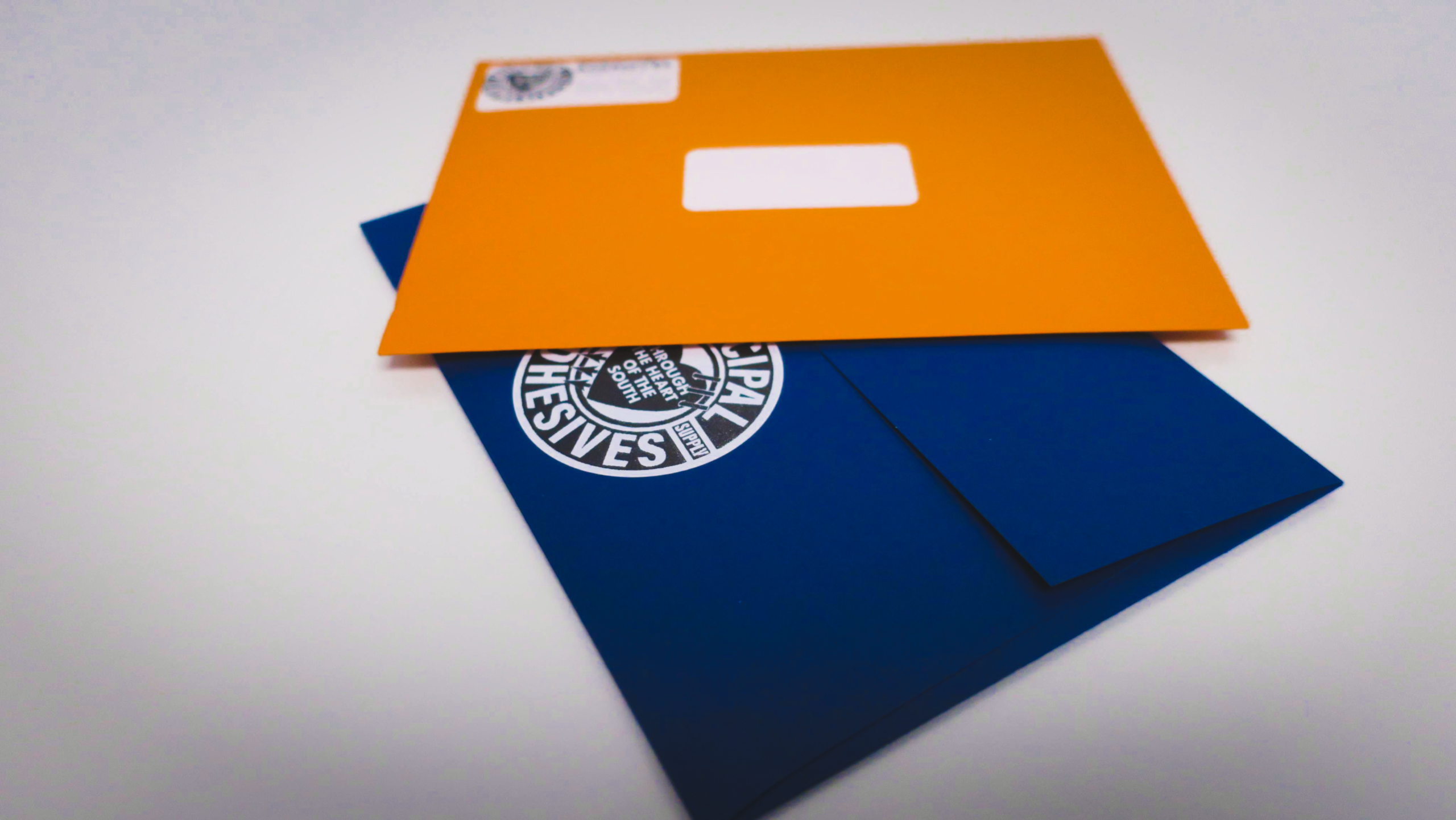 On a white smooth surface, blue and orange envelopes with Municipal Adhesives branding and return address.