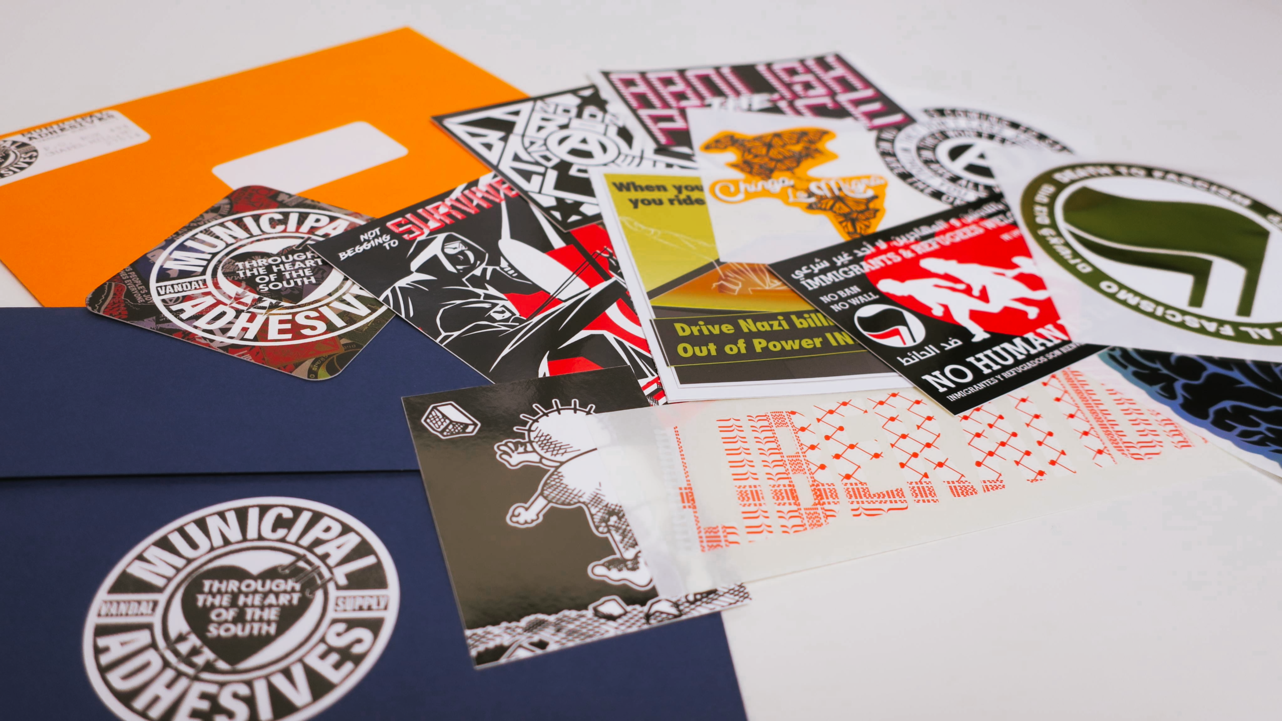 On a white smooth surface, a small envelope dumps some 12 Municipal Adhesives stickers along with a business card.