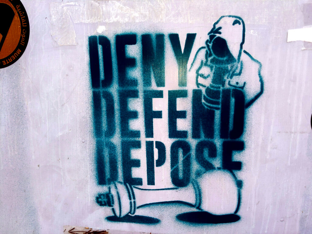 A closeup of a grey electrical box with a turquoise stencil painted reading Deny Defend Depose with a black pawn in a familiar hooded jacket and a fallen white king.