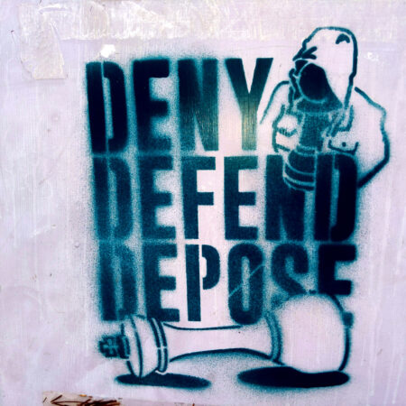 A closeup of a grey electrical box with a turquoise stencil painted reading Deny Defend Depose with a black pawn in a familiar hooded jacket and a fallen white king.