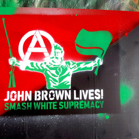 A green tarp with a black and red ascending diagonal corrugated plastic yard sign on top of it. Spraypainted on that is a simplified portrait of John Brown from John Steuart Curry's Tragic Prelude north wall mural. He holds black flags in his left hand and a rifle in his right. over his right shoulder, an anarchist circle-A. Below, the words "John Brown Lives! Smash White Supremacy"