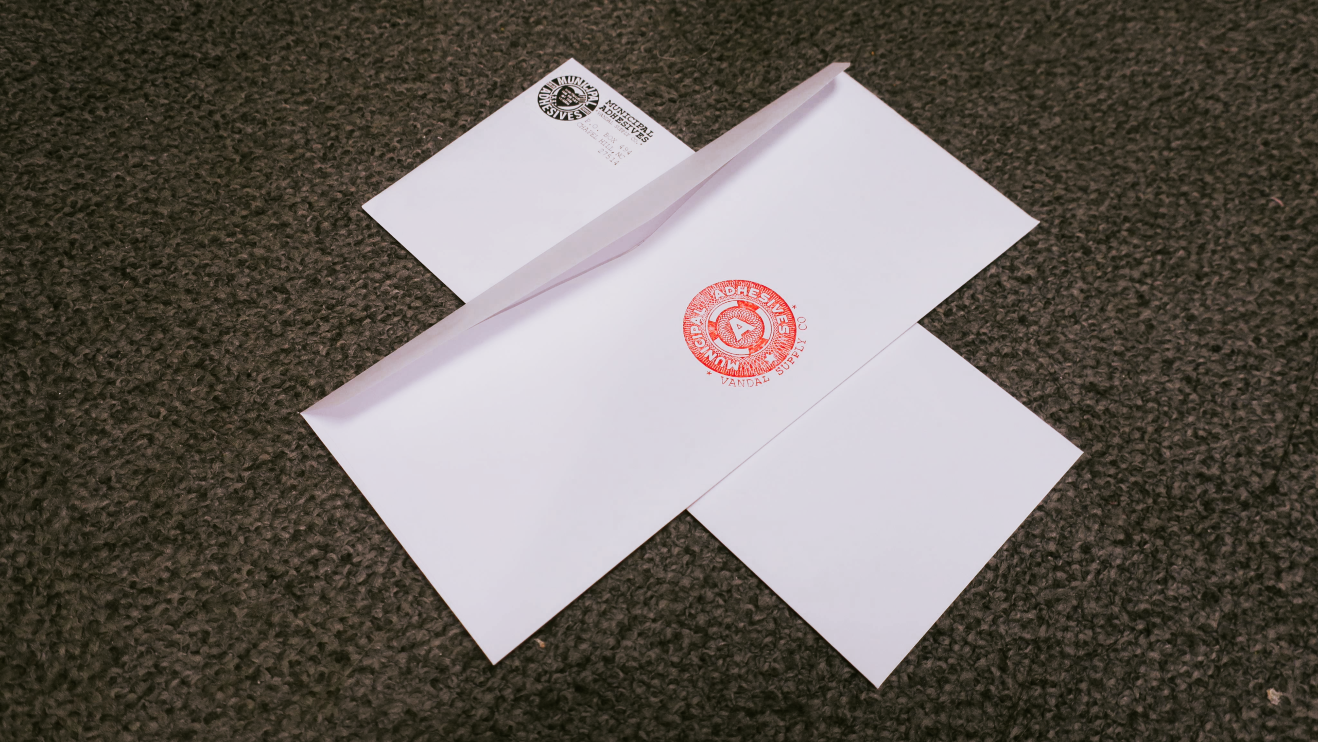 On a grey carpet, two crossed white standard envelopes with Municipal Adhesives branding and return addresses.
