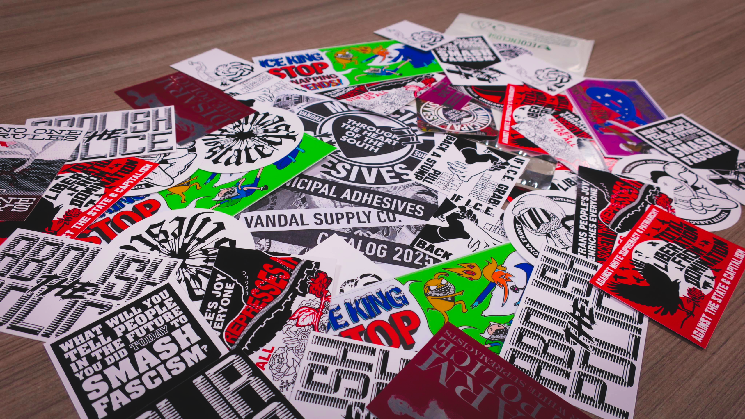 On a woodgrain surface, a polymailer envelope dumps some 40 Municipal Adhesives stickers along with a catalog and a business card.