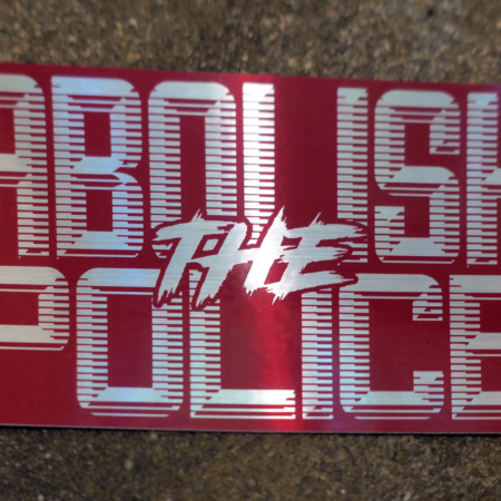 A concrete background with a rectangular sticker on it. The sticker is maroon, with brushed chrome letters in retrofuturistic typeface reading "Abolish the Police"