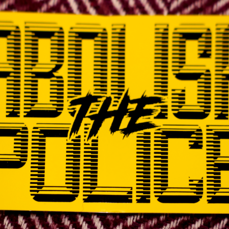 A purple and white wool chevron pattern with a rectangular sticker atop it. The sticker is bright yellow, with black retrofuturistic typeface reading "Abolish the Police"