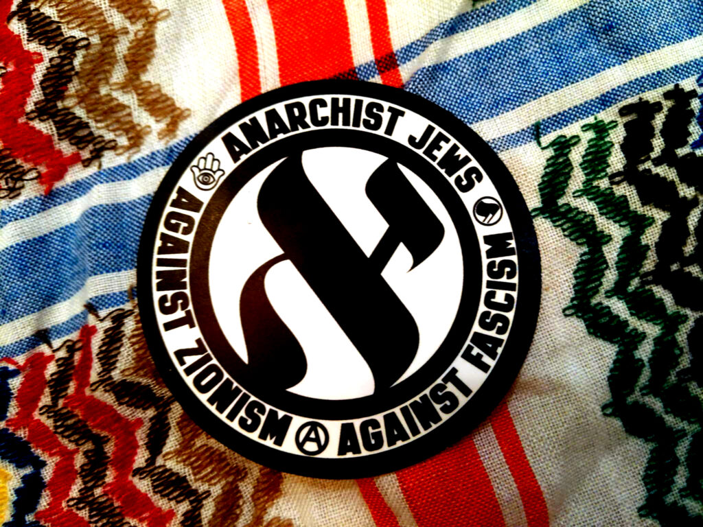 A rainbow-colored keffiyeh pattern with a circular sticker laid on top. A circled Aleph (first letter of "Anarchy" in Yiddish) surrounded by a circle of text reading "Anarchist Jews {Hamsa} Against Zionism {circle-A} Against Fascism {Antifa flags}."