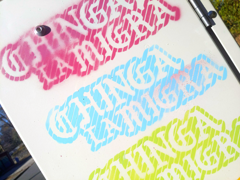 A close up of an electrical box with three colorful stenciled Chinga la Migra paintings, in pink, baby blue, and light green.