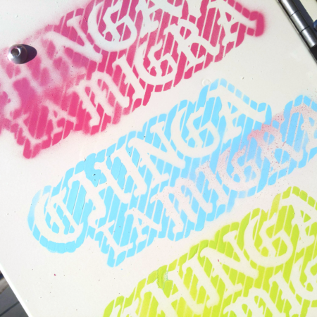 A close up of an electrical box with three colorful stenciled Chinga la Migra paintings, in pink, baby blue, and light green.