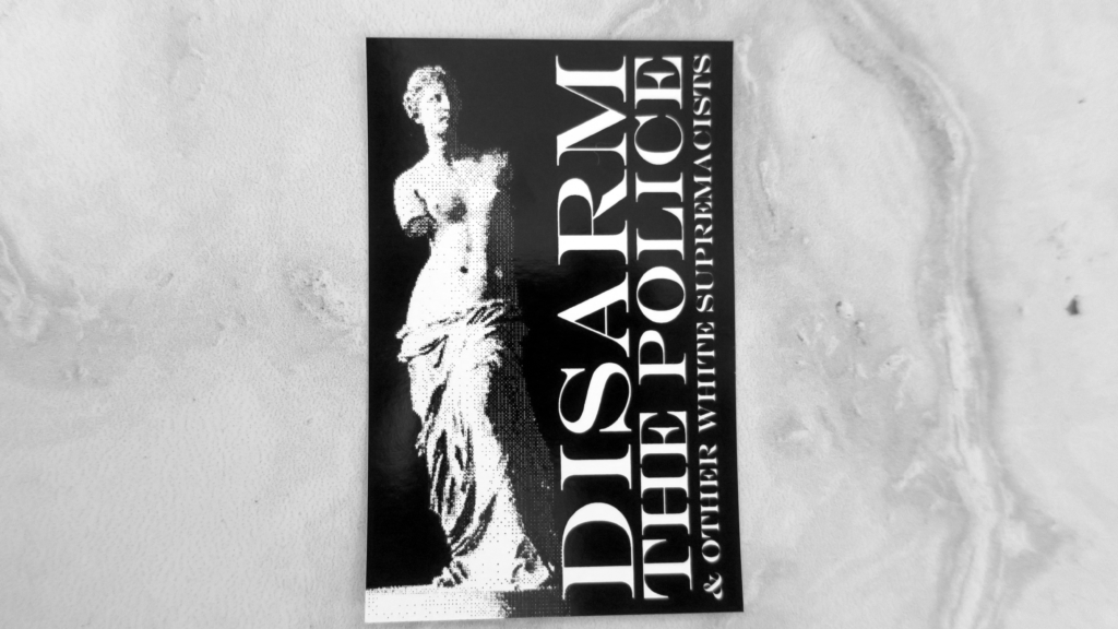 On a marbled surface, a black sticker with a white pixelated depiction of the Venus de Milo by sculptor Alexandros of Antioch. The words "Disarm the Police & Other White Supremacists" are tucked vertically facing upwards to the right of this statue in white.