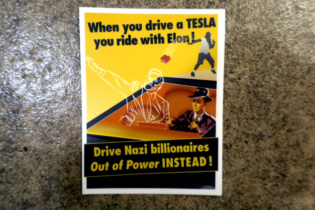 On a backdrop of concrete, a sticker: A white border around a yellow field with a cybertruck driving towards and past the viewer. The driver is wearing a purple suit and fedora. In the passenger seat of the cybertruck is an apparition of Elon Musk throwing a sieg heil salute. Behind him, a masked figure hurls a broken brick at the cybertruck. The words above the masked figure read "When you drive a TESLA you ride with Elon!" The words across the bottom of the sticker in a banner across the hood of the cybertruck read, "Drive Nazi billionaires Out of Power INSTEAD!"