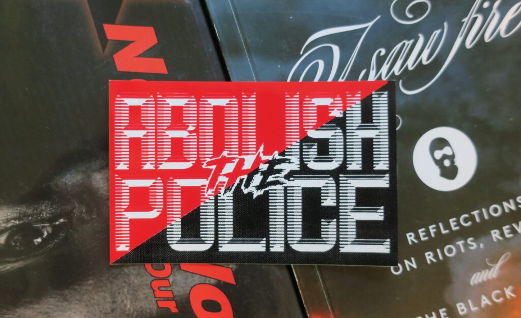 On a backdrop of anarchist books, a rectangular sticker: Ascending diagonal red and black with the words Abolish the Police printed in white with retro-futuristic type.