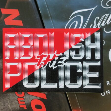 On a backdrop of anarchist books, a rectangular sticker: Ascending diagonal red and black with the words Abolish the Police printed in white with retro-futuristic type.