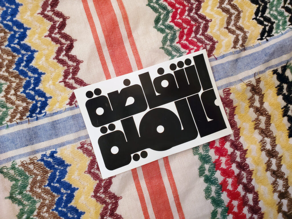 A rainbow keffiyeh pattern with a sticker on top of it. The stickers are black on white and read نتفاضة عالمية or Global Intifada in Arabic in a round 70s-looking script.