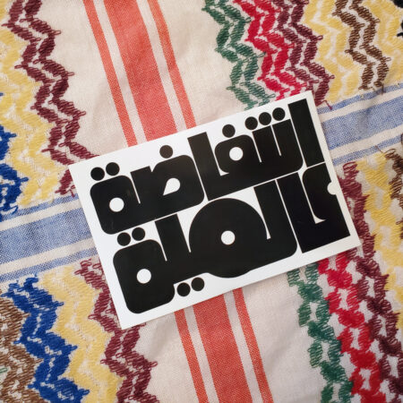 A rainbow keffiyeh pattern with a sticker on top of it. The stickers are black on white and read نتفاضة عالمية or Global Intifada in Arabic in a round 70s-looking script.