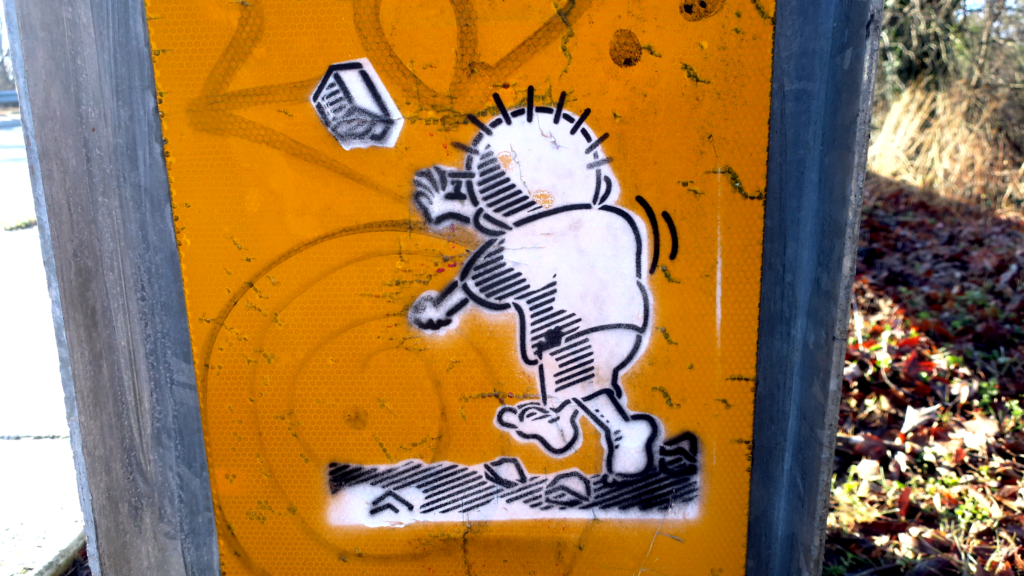 A photograph of the end guard of a traffic guardrail. Over the grimy and tagged yellow reflective surface a two layer stencil painting of a cartoon of a shoeless boy in tattered clothing (Handala) facing away from the viewer, launching a rock into the air at an unseen oppressor.