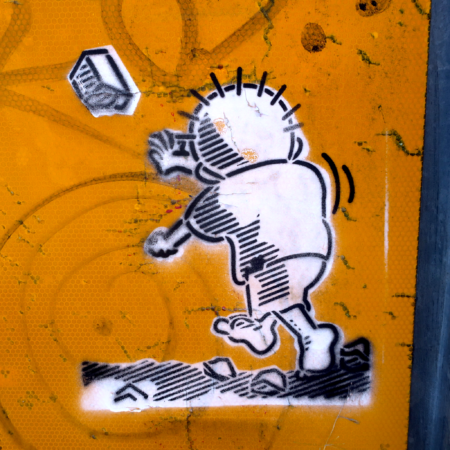 A photograph of the end guard of a traffic guardrail. Over the grimy and tagged yellow reflective surface a two layer stencil painting of a cartoon of a shoeless boy in tattered clothing (Handala) facing away from the viewer, launching a rock into the air at an unseen oppressor.