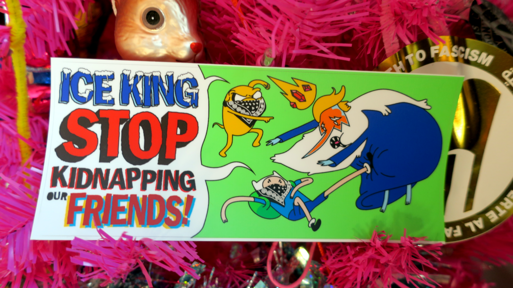 Inside a pink pine tree with a gold Death to Fascism sticker, a gilded squirrel and some holographic tinsel hangs a green sticker. On the sticker, Jake the Dog and Finn the human, inexpertly bloc'd up with black bandanas, yell at the Ice King "Stop Kidnapping our FRIENDS!" Ice King is wearing spray tan on his face and has a blonde swoop hairdo, making him the ICE king, though his crown was knocked off by a swift kick to the groin from Finn the human.