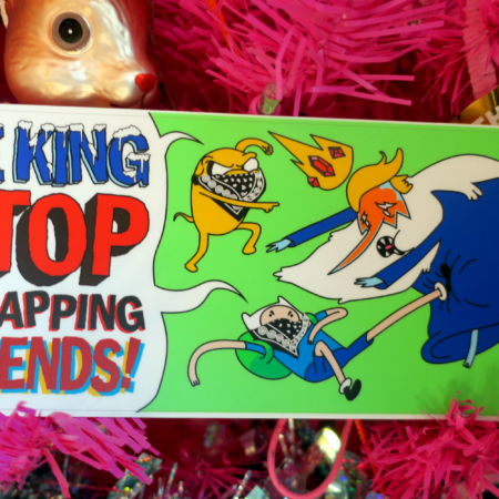 Inside a pink pine tree with a gold Death to Fascism sticker, a gilded squirrel and some holographic tinsel hangs a green sticker. On the sticker, Jake the Dog and Finn the human, inexpertly bloc'd up with black bandanas, yell at the Ice King "Stop Kidnapping our FRIENDS!" Ice King is wearing spray tan on his face and has a blonde swoop hairdo, making him the ICE king, though his crown was knocked off by a swift kick to the groin from Finn the human.