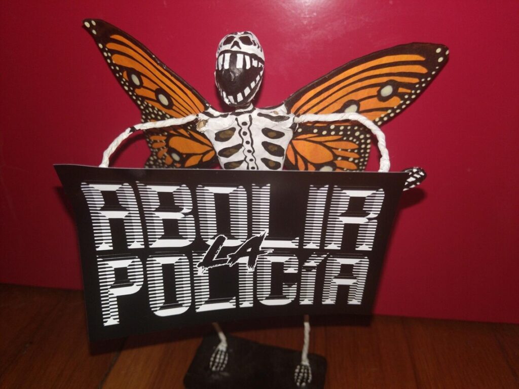 Un esqueleto con alas de mariposa sosteniendo una etiqueta que dice "Abolir la policía" en tipografía retrofuturista blanca sobre fondo negro.