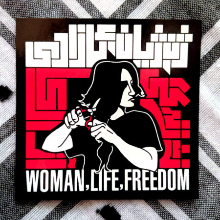 A sticker on a keffiyeh: A woman cutting her hair off, a Persian symbol of mourning or protest. She is flanked by geometric kufic Persian script in red, topped by geometric kufic Arabic Kurmanji script in white, with English words below, all reading, "Woman, Life, Freedom"'