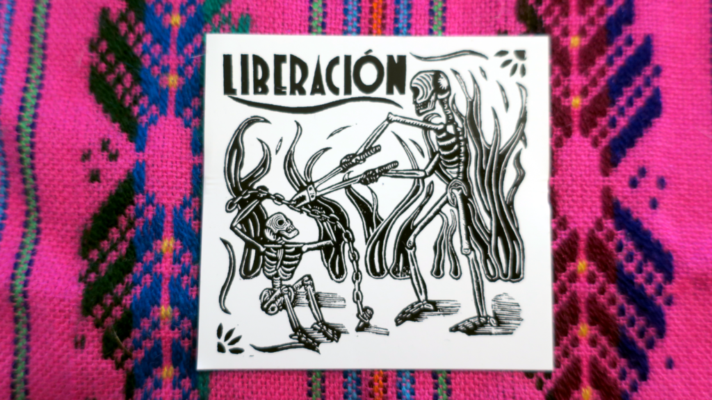 On a colorful pink embroidered tapestry, a white sticker: A woodcut illustration of two skeletons in the midst of a fire, one of whom is using a pair of large boltcutters to unchain a second skeleton. The word Liberación, Spanish for Liberation, appears above in an art-deco style type.