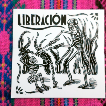On a colorful pink embroidered tapestry, a white sticker: A woodcut illustration of two skeletons in the midst of a fire, one of whom is using a pair of large boltcutters to unchain a second skeleton. The word Liberación, Spanish for Liberation, appears above in an art-deco style type.