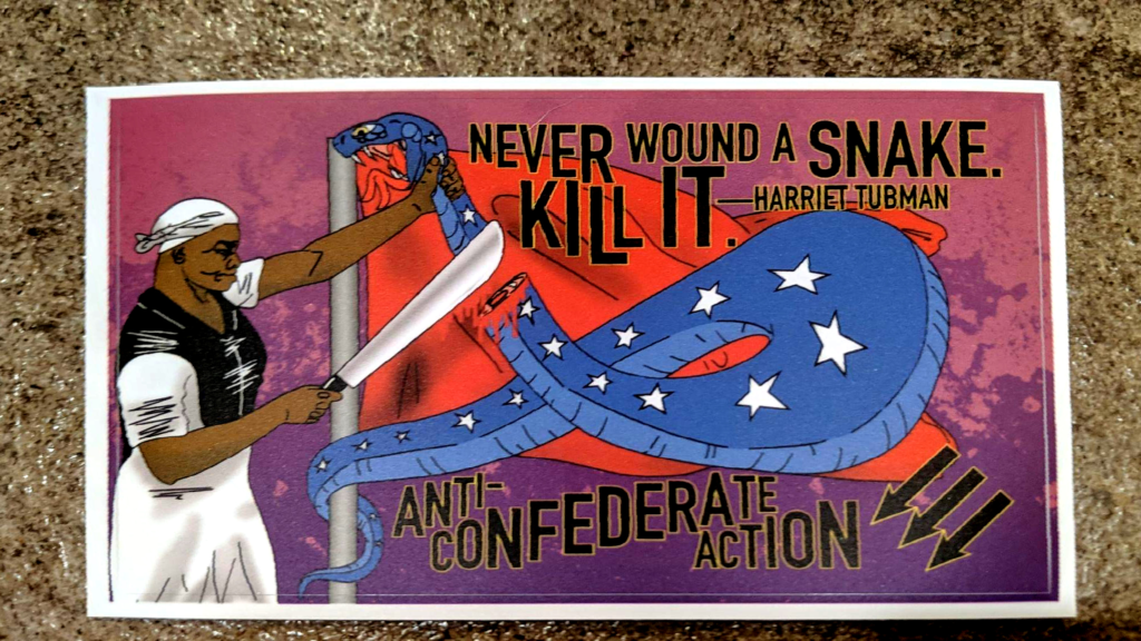 On a background of concrete, an Anti-Confederate Action sticker: Harriet Tubman stands mean-mugging a snake she has severed the head from with a machete. The snake is curled and doubled over on itself, having been unraveled from a flagpole upon which is a red flag, giving the impression of a confederate battle flag. The quotation, "Never wound a snake. Kill it. - Harriet Tubman" appears above the flag. The words, "Anti-Confederate Action" appear below in black with yellow border. There are three antifascist arrows in the same colors in the bottom right corner.