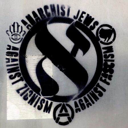 A metal sign has a stencil spray painted on it: A circled aleph. The words "Anarchist Jews Against Zionism Against Fascism" wrap around the circle. Between these phrases, a hamsa (a symbol inclusive of Jewish culture not associated with the state of Israel), a circle-A, and antifascist flag logo.