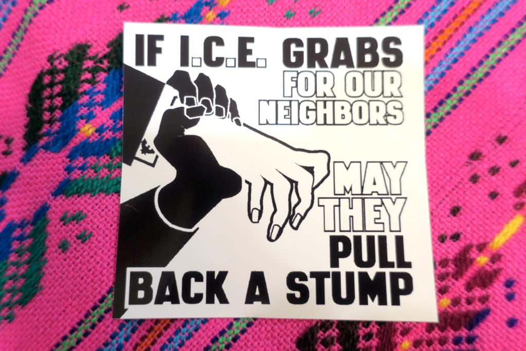 On a pink colorful tapestry, a black and white Municipal Adhesives sticker: a thin white hand in a dark suit with a white exposed cuff that bears a Department of Homeland Security cufflink reaches out but is gripped by a darker hand, stopping it dead. Words reading : "If I.C.E. Grabs for Our Neighbors May They Pull Back a Stump" appear to the right of the two hands.