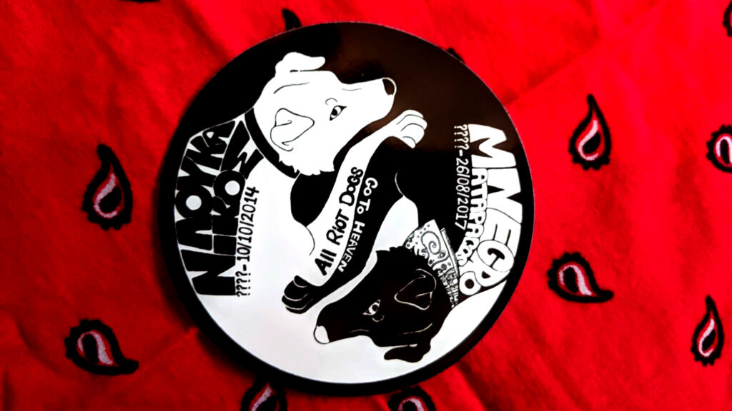 A background of a red paisley bandana with a sticker in the shape of a circle: a black and white portrait of two intertwined dogs, one black and one white. The white one wears a collar, the black one wears a bandana. Across their paws are the words "All riot dogs go to heaven." On the back of the white dog, the words "Λουκάνικος (????-10/10/2014)." On the back of the black dog, the words "Negro Matapacos (????-26/08/2017)"