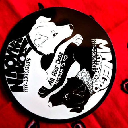 A background of a red paisley bandana with a sticker in the shape of a circle: a black and white portrait of two intertwined dogs, one black and one white. The white one wears a collar, the black one wears a bandana. Across their paws are the words "All riot dogs go to heaven." On the back of the white dog, the words "Λουκάνικος (????-10/10/2014)." On the back of the black dog, the words "Negro Matapacos (????-26/08/2017)"