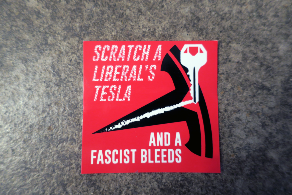 On top of smooth concrete, a red sticker with "Scratch a liberal's Tesla and a Fascist Bleeds." printed on it. This message is bisected by a key dragging a scrape across Tesla's logo.