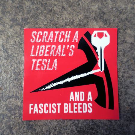 On top of smooth concrete, a red sticker with "Scratch a liberal's Tesla and a Fascist Bleeds." printed on it. This message is bisected by a key dragging a scrape across Tesla's logo.