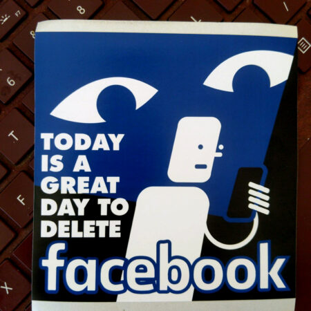 A maroon keyboard with a sticker that reads "Today is a Good Day to Delete Facebook" with an image of someone basked in the blue light of their phone which projects large surveilling eyes onto the wall in back of them.