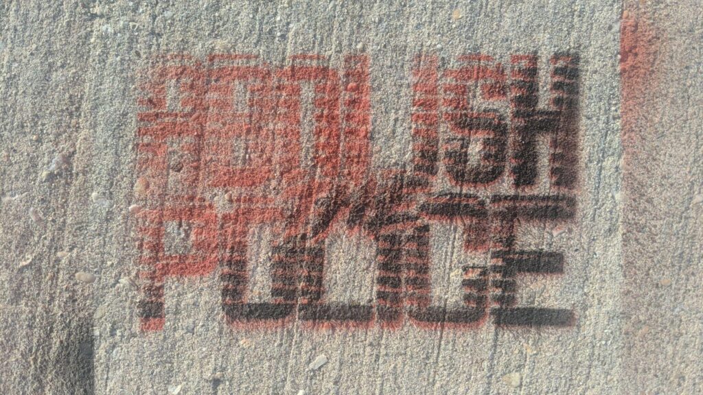 A rough concrete surface spraypainted through a stencil "Abolish the Police" in red and black ascending diagonal.