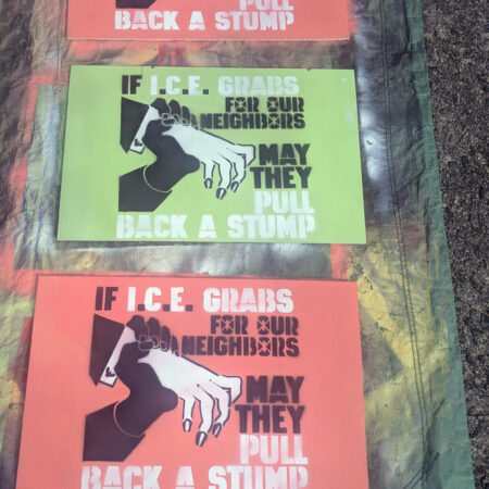 On a green tarp, three spraypainted corrugated plastic signs. Two have a salmon colored background, and the middle one has a chartreuse background. On these signs is painted a thin white hand in a dark suit with a white exposed cuff that bears a Department of Homeland Security cufflink reaches out but is gripped by a darker hand, stopping it dead. Words reading "If I.C.E. Grabs for Our Neighbors May They Pull Back a Stump" appear to the right of the two hands.