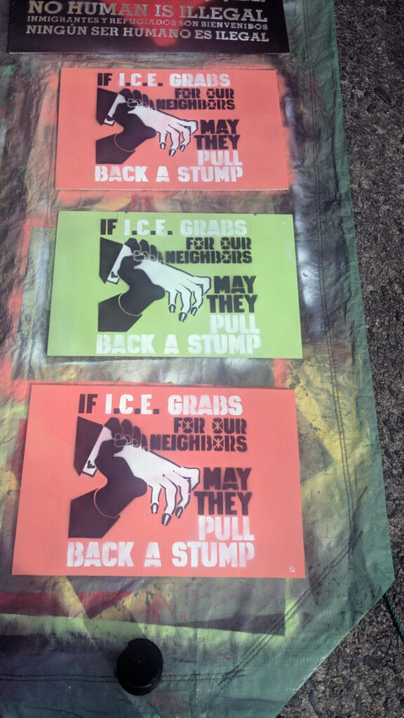 On a green tarp, three spraypainted corrugated plastic signs. Two have a salmon colored background, and the middle one has a chartreuse background. On these signs is painted a thin white hand in a dark suit with a white exposed cuff that bears a Department of Homeland Security cufflink reaches out but is gripped by a darker hand, stopping it dead. Words reading "If I.C.E. Grabs for Our Neighbors May They Pull Back a Stump" appear to the right of the two hands.