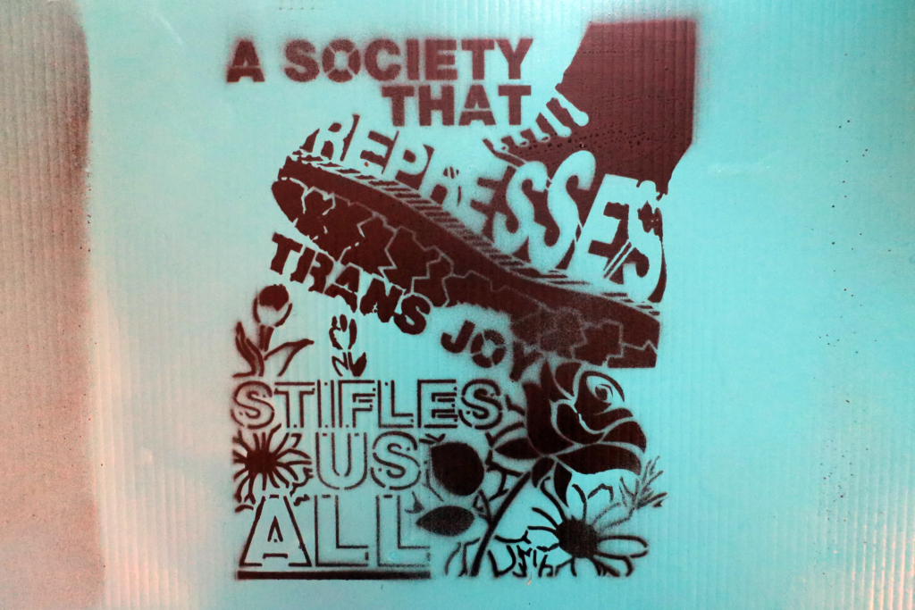 A blue corrugated plastic surface spray painted with a black stencil that reads: "A society that represses trans joy stifles us all" These words are twisted around and inside of a boot that appears to step on a rose, but which will actually trample a whole garden of flowers.