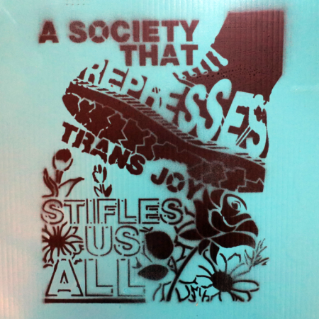 A blue corrugated plastic surface spray painted with a black stencil that reads: "A society that represses trans joy stifles us all" These words are twisted around and inside of a boot that appears to step on a rose, but which will actually trample a whole garden of flowers.