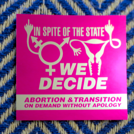 On a blue and white chevron pattern, a sticker in rhodamine red on mirror chrome: a uterus whose lateral Fallopian tube is twisted up and folded into a middle finger gesture, and a trans symbol similarly flipping the bird. Between these symbols, the words "In Spite of the State We Decide; Abortion & Transition on Demand Without Apology" appear.