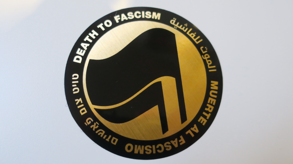 On a blank background, a sticker: A thick black circle with the words: "Death to Fascism, Muerte al Fascismo, الموت للفاشية, טויט צום פֿאַשיזם, ." Inside this circle are two black flags arranged over gold chrome recognizable as the antifascist logo.