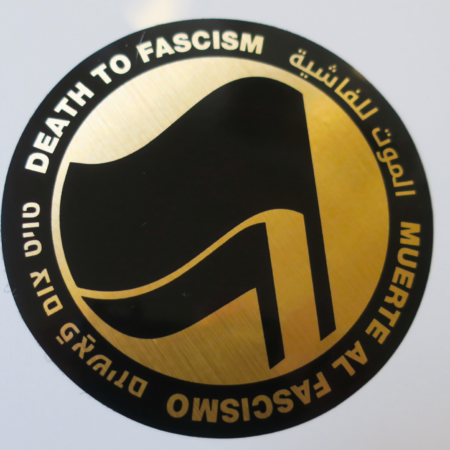 On a blank background, a sticker: A thick black circle with the words: "Death to Fascism, Muerte al Fascismo, الموت للفاشية, טויט צום פֿאַשיזם, ." Inside this circle are two black flags arranged over gold chrome recognizable as the antifascist logo.