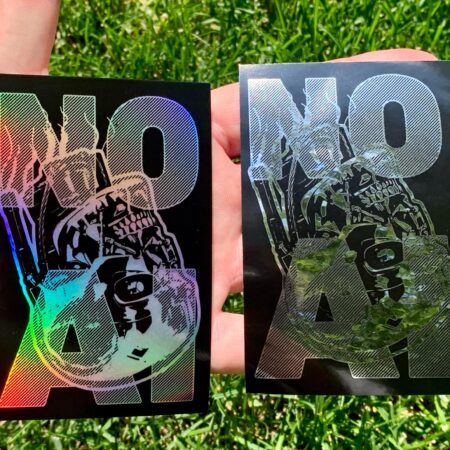Against a background of grass, a hand holds two stickers on its open palm, the first on the left is a black sticker with rainbow holograph, the second on the right with mirror chrome. Both stickers display a severed terminator head overlaid with the words "NO AI."