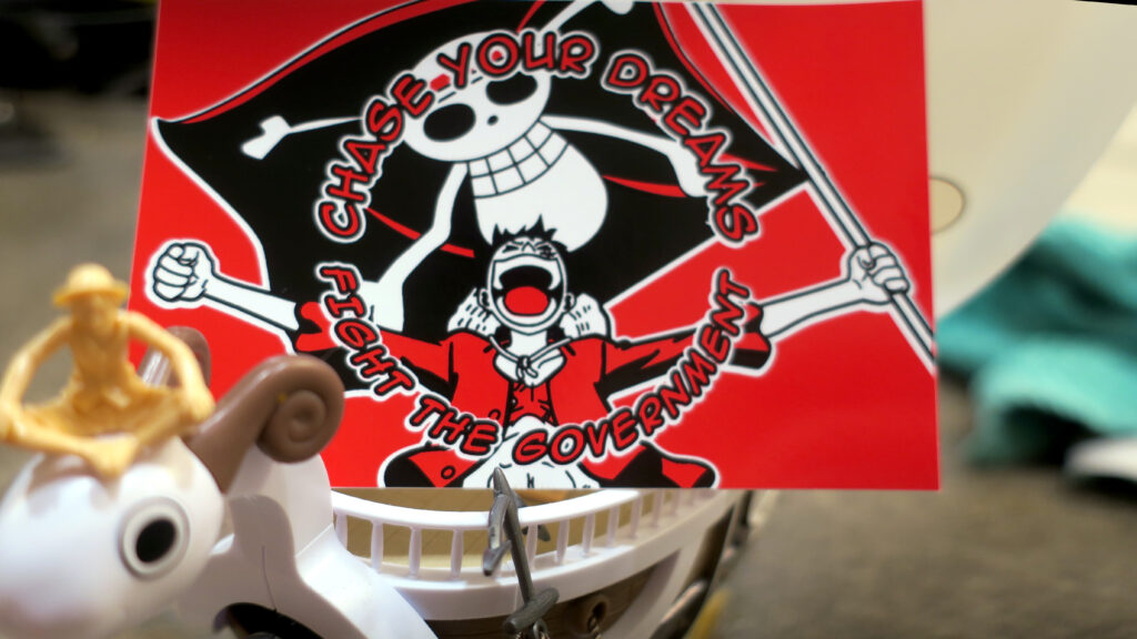 A red and black sticker perched on the deck of the Going Merry with Luffy sitting atop the goat figurehead. The sticker reads "Chase Your Dreams//Fight the Government" in comic book lettering arcing above and below a beloved fighter for freedom and friendship who holds a huge black flag emblazoned with Gen Z's protest image.