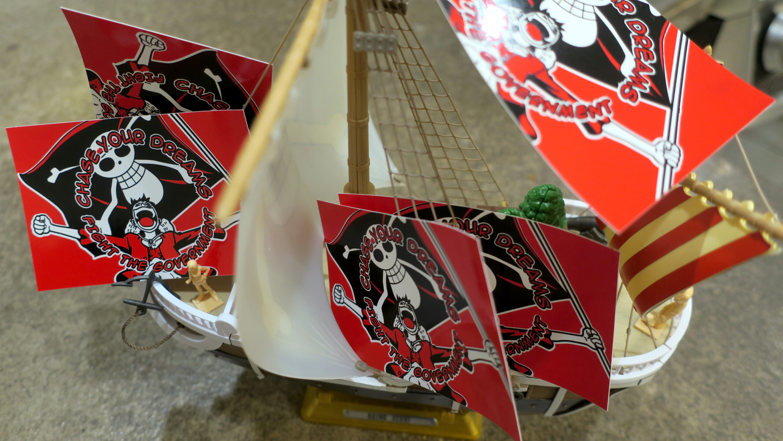 The Going Merry sits on a concrete background, red black and white stickers adorn its rigging, deck and the lateen sail. The stickers are red black and white, and it reads "Chase Your Dreams//Fight the Government" in comic book lettering arcing above and below a beloved fighter for freedom and friendship who holds a huge black flag emblazoned with Gen Z's protest image.
