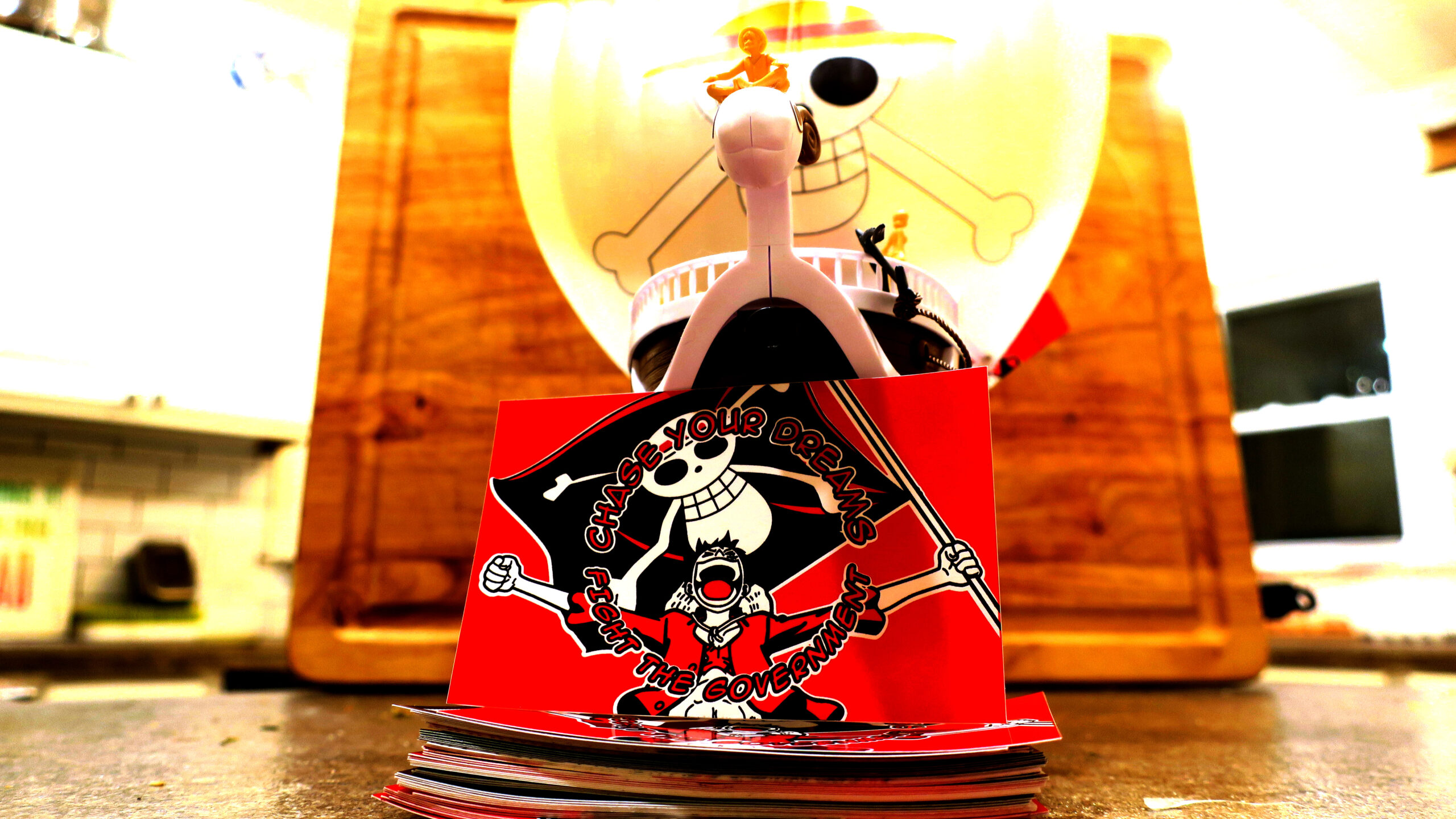On a woodgrain background, the Going Merry towers with Luffy sitting atop the goat figurehead. The ship towers over a tidy stack of red black and white stickers, the top of which is erected toward the viewer: It reads "Chase Your Dreams//Fight the Government" in comic book lettering arcing above and below a beloved fighter for freedom and friendship who holds a huge black flag emblazoned with Gen Z's protest image.