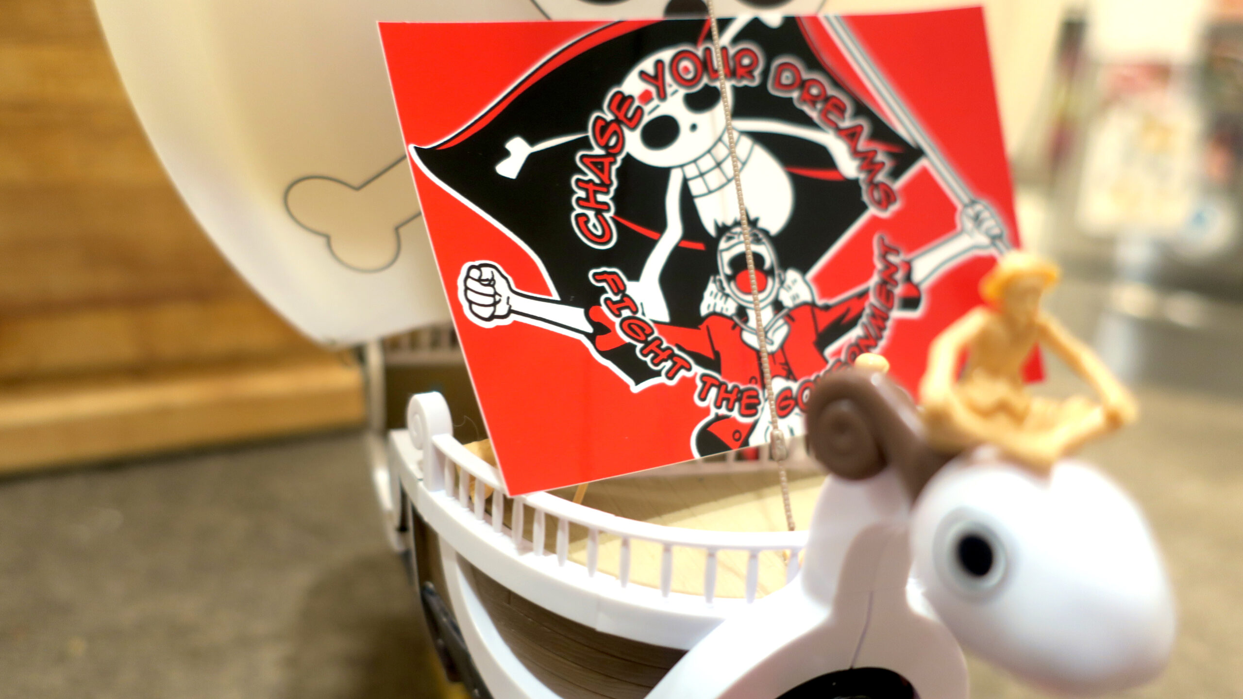 A red and black sticker perched on the deck of the Going Merry with Luffy sitting atop the goat figurehead. The sticker reads "Chase Your Dreams//Fight the Government" in comic book lettering arcing above and below a beloved fighter for freedom and friendship who holds a huge black flag emblazoned with Gen Z's protest image.